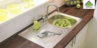 Νεροχύτης αλουμινίου με διάφορα μαγειρικά σκεύη, λαχανικά και φρούτα, πάνω σε καφέ πάγκο κουζίνας